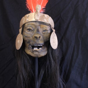 May include: A detailed, realistic human skull mask with a gold headdress and red feathers. The mask has a painted expression of surprise and is mounted on a black stand.