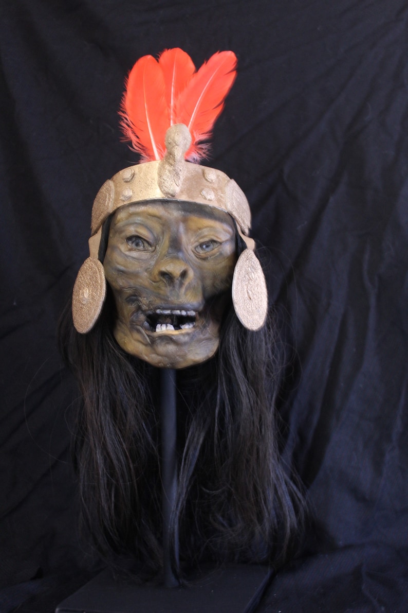 May include: A detailed, realistic human skull mask with a gold headdress and red feathers. The mask has a painted expression of surprise and is mounted on a black stand.