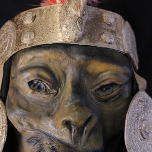 May include: A close-up of a detailed, realistic, sculpted mask with a fierce expression. The mask is painted in shades of brown and green, with a large, open mouth revealing teeth. The mask is adorned with a gold headband and a red feather headdress.