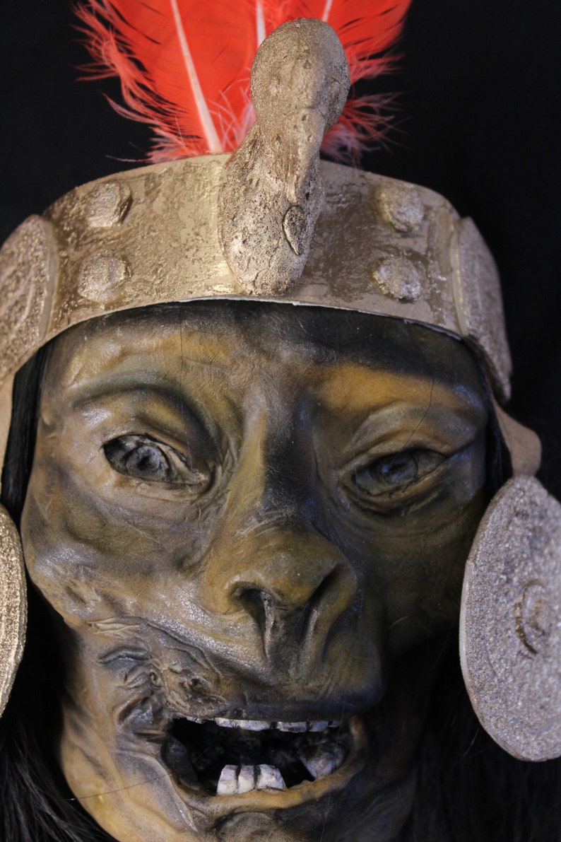 May include: A close-up of a detailed, realistic, sculpted mask with a fierce expression. The mask is painted in shades of brown and green, with a large, open mouth revealing teeth. The mask is adorned with a gold headband and a red feather headdress.