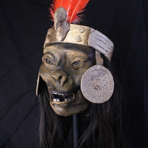 May include: A green and brown mask with a toothy grin, wearing a gold and silver headdress with red feathers. The mask has a long black wig.