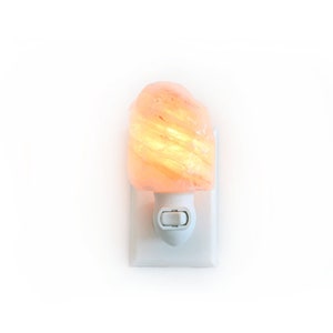 Può includere: Una luce notturna al sale dell'Himalaya. La lampada ha un cristallo di sale grezzo, arancione-rosa, sopra una base bianca a spina. Il cristallo di sale si illumina con una luce calda e gialla quando è illuminato.