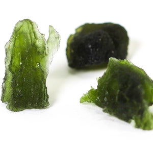 May include: Three pieces of raw, translucent green moldavite gemstones. The gemstones have irregular shapes and a textured surface, set against a white background. The color is a deep, vibrant green.