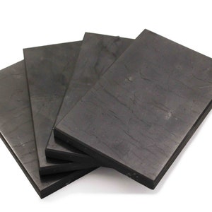 May include: Four rectangular, dark gray slate tiles are stacked slightly, revealing their textured surfaces. The tiles have a natural, stone-like appearance, with subtle variations in color and veining. The edges are clean and defined.