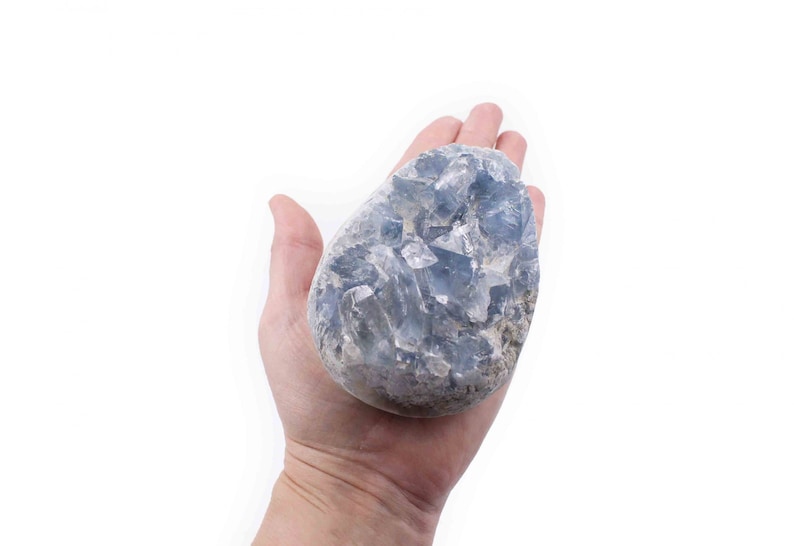 May include: A large, light blue crystal cluster with a rough, natural texture. The crystal is held in a hand, showcasing its size and shape.