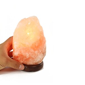 May include: A pink Himalayan salt lamp with a warm, orange glow. The lamp is on a wooden base.