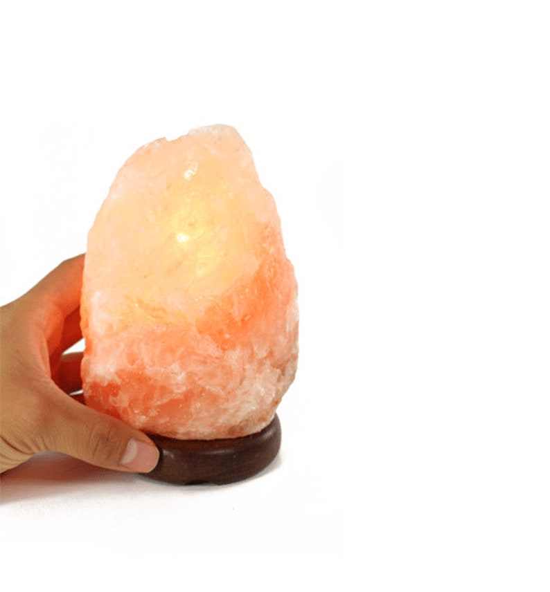 May include: A pink Himalayan salt lamp with a warm, orange glow. The lamp is on a wooden base.