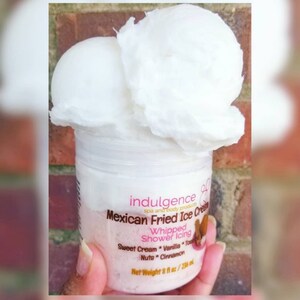 Pu&ograve; includere: Un barattolo bianco con un'etichetta rosa che recita "indulgence spa and body products Mexican Fried Ice Cream Whipped Shower Icing Sweet Cream Vanilla Toppings Nuts Cinnamon Net Weight 8 fl oz / 236 ml". Il barattolo &egrave; sormontato da due cumuli di panna montata bianca.
