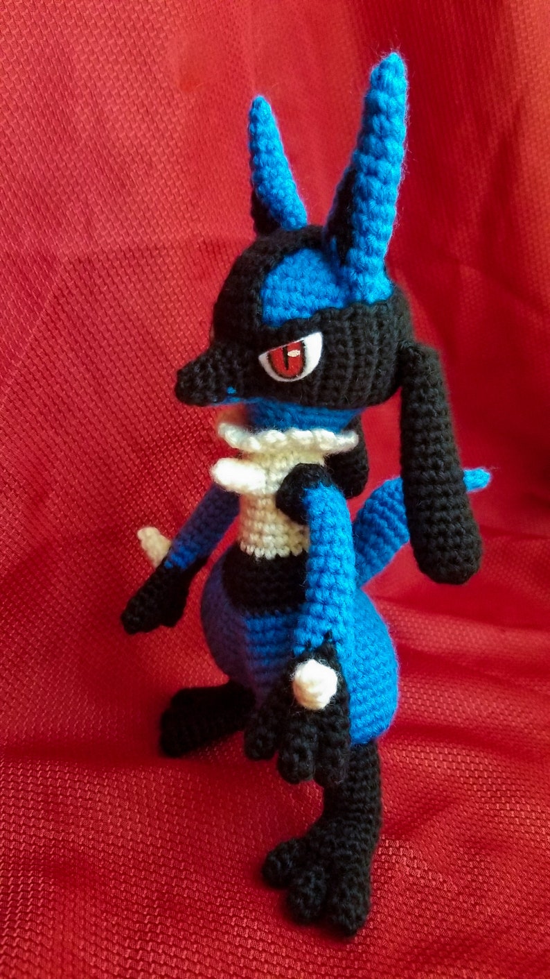 Puede incluir: Un juguete de peluche azul y negro de crochet de un personaje de Pokemon. El juguete tiene un cuello blanco y ojos rojos.