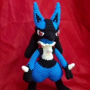 Puede incluir: Un juguete de peluche azul y negro de crochet de un personaje de Pokemon con ojos rojos. El juguete est&aacute; de pie sobre una superficie roja.