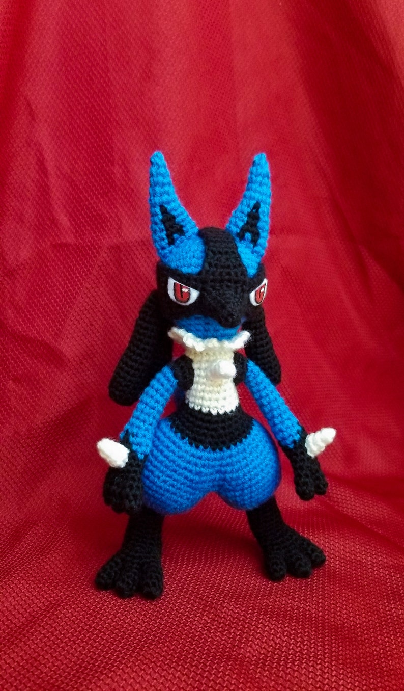 Puede incluir: Un juguete de peluche azul y negro de crochet de un personaje de Pokemon con ojos rojos. El juguete est&aacute; de pie sobre una superficie roja.