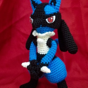 Puede incluir: Un juguete de peluche azul y negro tejido a crochet con ojos rojos. El juguete est&aacute; de pie sobre una superficie roja.