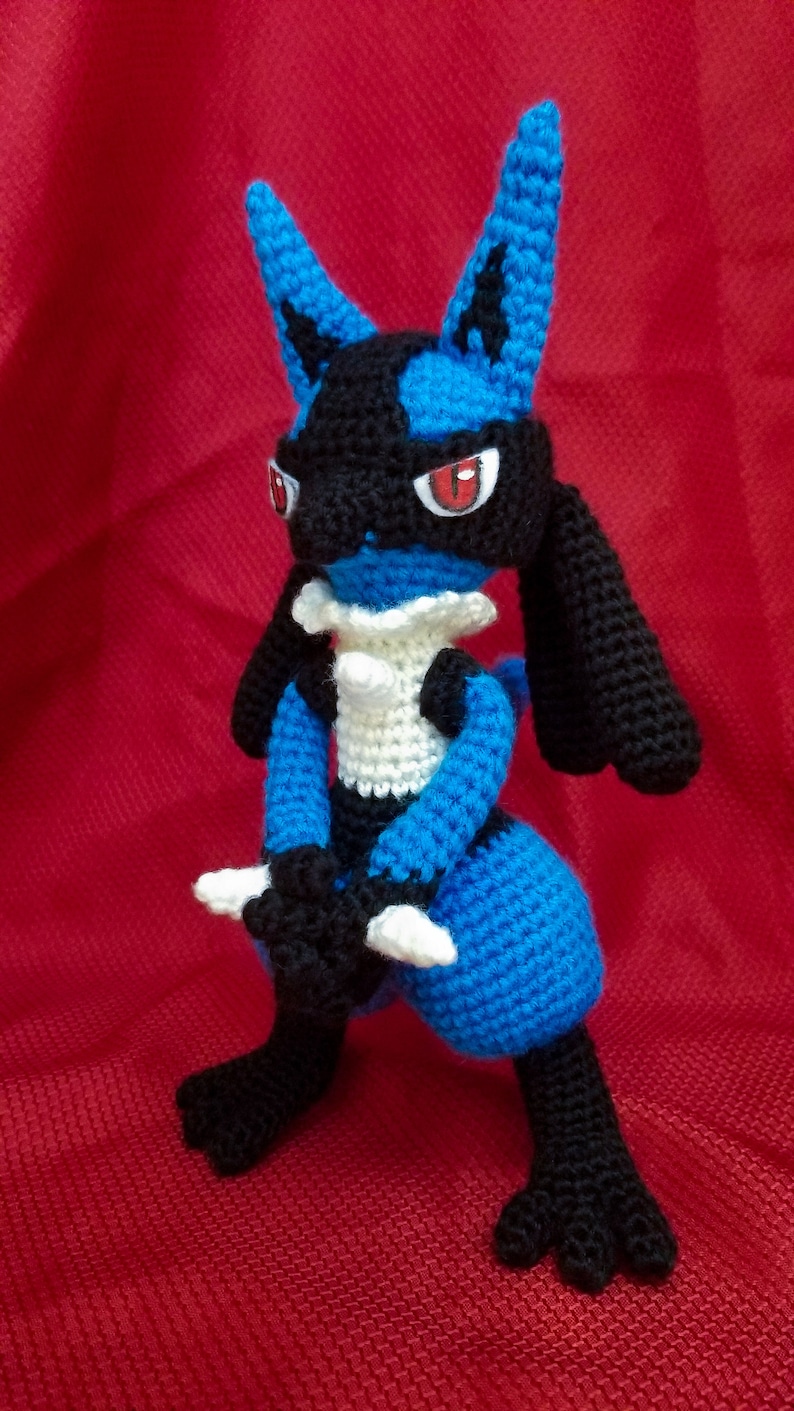 Puede incluir: Un juguete de peluche azul y negro tejido a crochet con ojos rojos. El juguete est&aacute; de pie sobre una superficie roja.