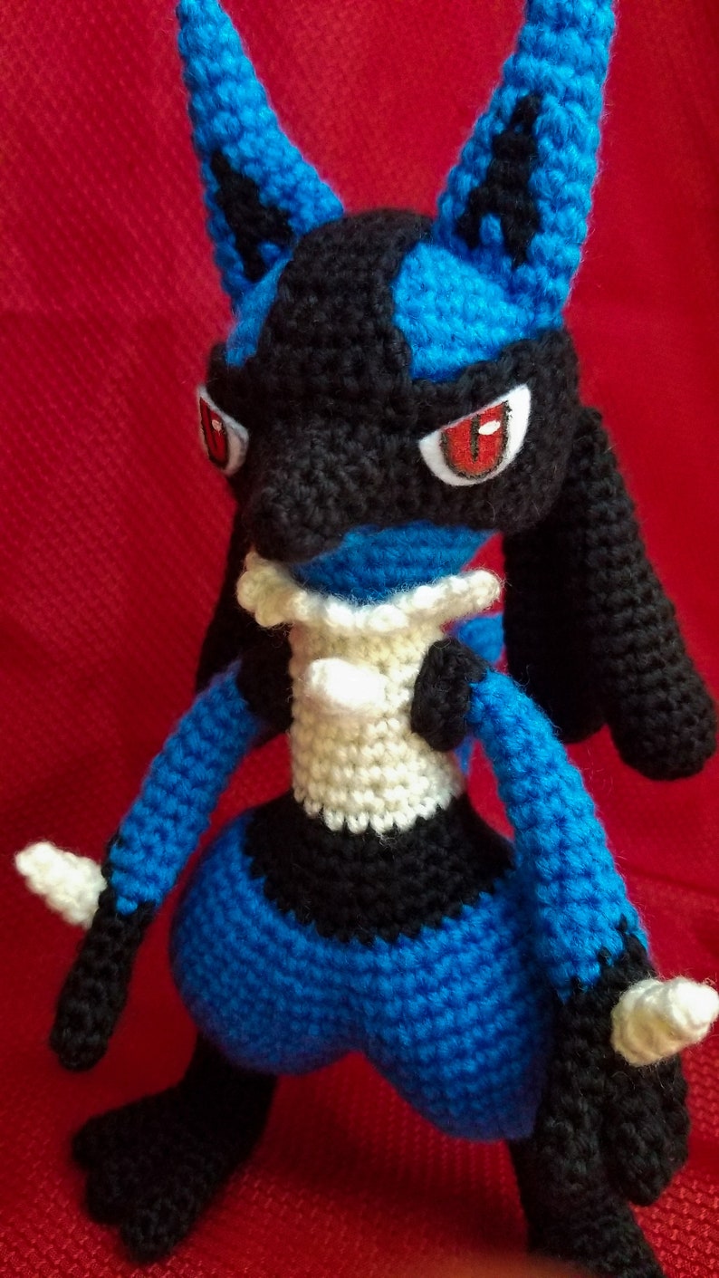 Puede incluir: Un juguete de peluche azul y negro tejido a crochet con ojos rojos. El juguete es un personaje popular de un videojuego y una serie de anime.