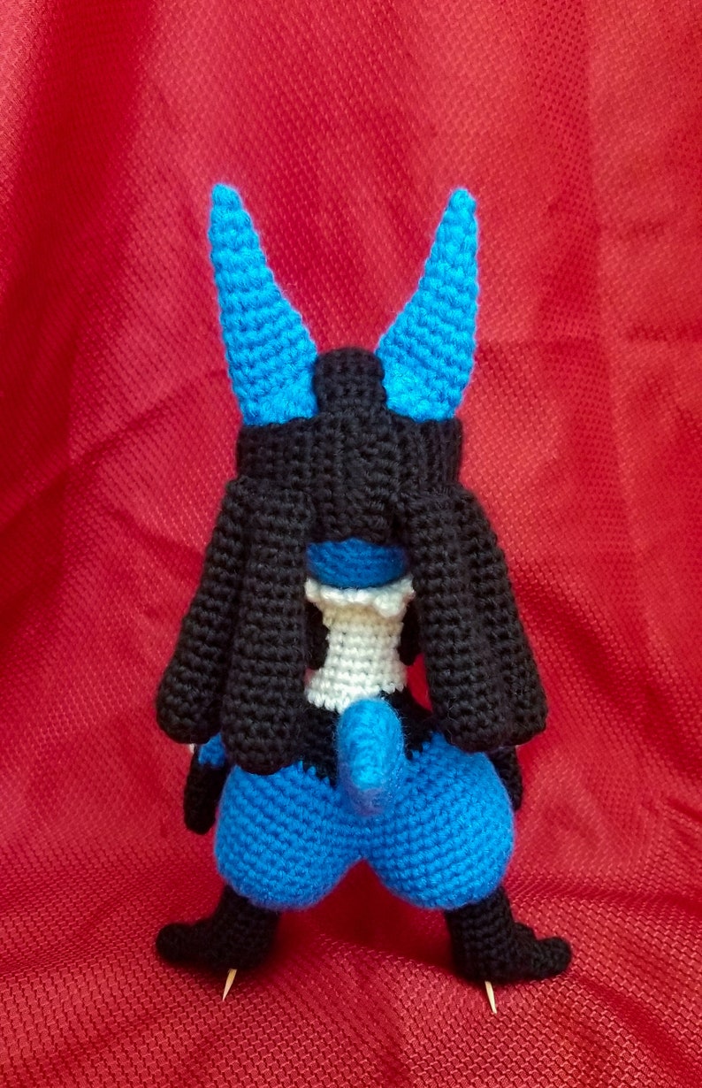 Puede incluir: Un juguete de peluche azul y negro tejido a crochet con una cola larga y orejas puntiagudas. El juguete est&aacute; de pie sobre una superficie roja.