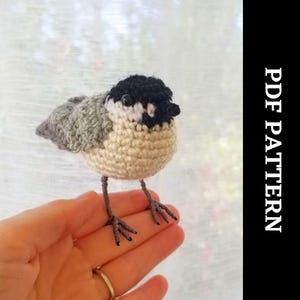 Könnte beinhalten: Häkelanleitung für einen kleinen Vogel mit schwarzen Augen und einem schwarzen Kopf. Der Vogel ist in beige und grauem Garn gehäkelt und hat einen kleinen Schnabel. Der Vogel sitzt auf einer Hand.