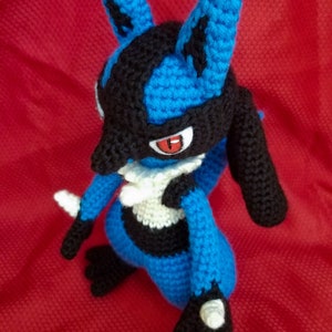 Puede incluir: Un juguete de peluche azul y negro tejido a crochet con un pecho blanco y ojos rojos. El juguete est&aacute; de pie sobre una superficie roja.