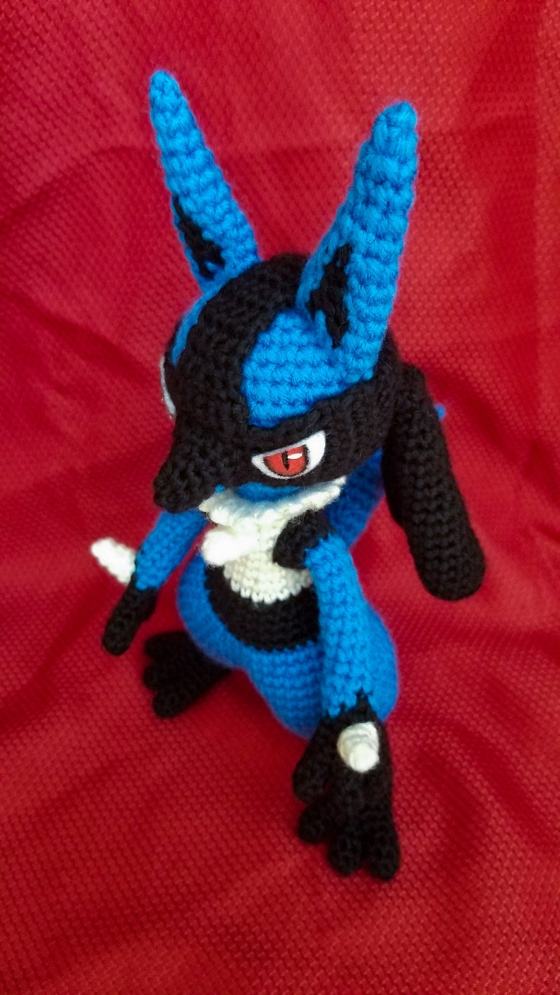 Puede incluir: Un juguete de peluche azul y negro tejido a crochet con un pecho blanco y ojos rojos. El juguete est&aacute; de pie sobre una superficie roja.