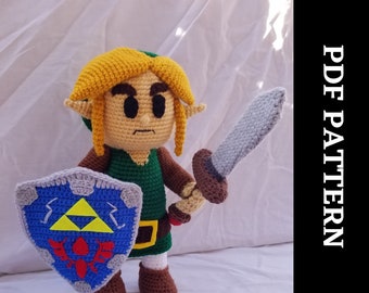 May include: Crocheted Link plush toy holding a sword and shield. The plush is green, brown, and yellow. The shield is blue and gray with a yellow triangle design. The sword is gray with a brown handle.