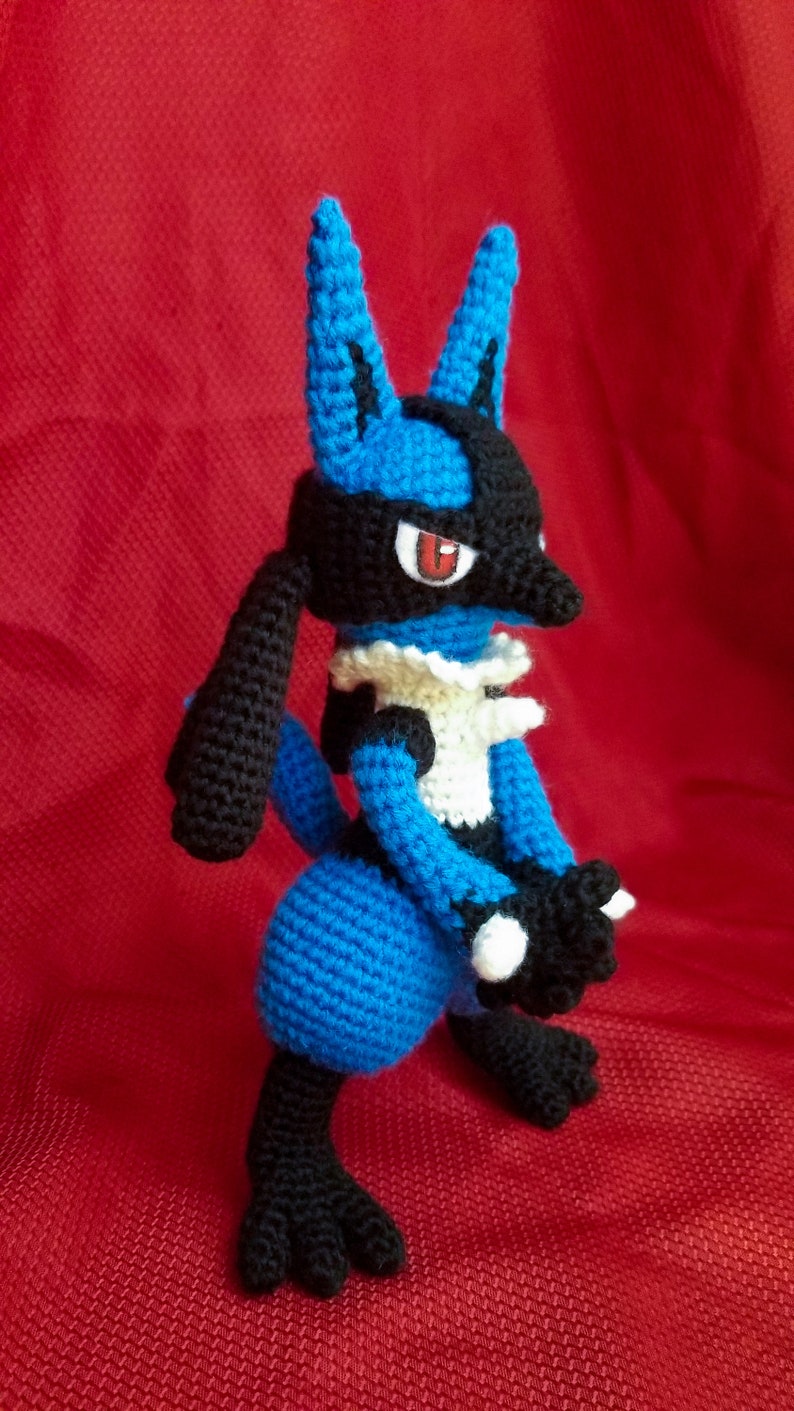 Puede incluir: Un juguete de peluche azul y negro de crochet de un personaje de Pokemon. El juguete tiene un cuello blanco y ojos rojos.