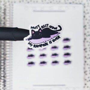 May include: A sticker featuring a black cat resting on a purple pillow. The text "MUST REST NOW MY RAMPAGE IS OVER" is written above and below the cat. Additional stickers with purple accents are visible.