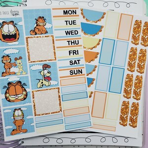 May include: Planner stickers featuring Garfield and Odie characters. The stickers include weekly layouts, decorative elements, and glitter accents. The color palette includes orange, blue, and gold, with the days of the week clearly labeled.