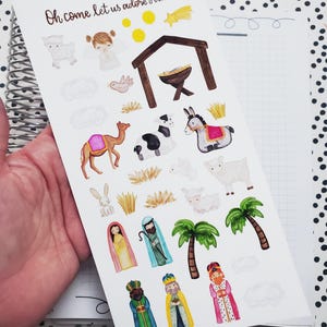 May include: A sheet of Christmas stickers featuring a nativity scene with a manger, a donkey, a camel, a cow, sheep, angels, a star, palm trees, and the three wise men. The text "Oh come let us adore Him" is at the top of the sheet.