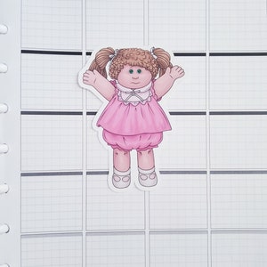 May include: A pink and white doll sticker with brown hair and pigtails. The doll is wearing a pink dress with a white collar and white shoes.