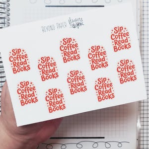 May include: A sheet of white stickers with the text "Sip Coffee Read Books" in red. The stickers are oval-shaped and feature a starburst design. The sheet is held over a notebook with a grid pattern.