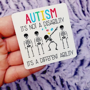 May include: A white sticker with the text "AUTISM IT'S NOT A DISABILITY IT'S A DIFFERENT ABILITY" in colorful letters. Four black skeleton figures are shown, one of them is doing a dabbing pose. There are small colorful puzzle pieces scattered around the skeletons.