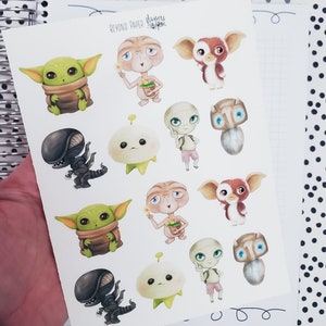 May include: A sheet of stickers featuring various characters from popular movies, including ET, the alien from ET the Extra-Terrestrial, a creature from the Alien franchise, and a character from the movie Gremlins. The stickers are on a white background and are printed with vibrant colors.