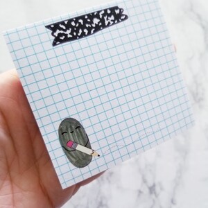 May include: A white gridded notepad with a black and white patterned tape at the top. A small black and gray cartoon character is holding a pencil and drawing a dotted line.