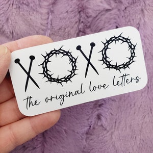 May include: White sticker with black text and graphics. The text reads "XOXO the original love letters". The graphics are two crossed needles and a crown of thorns.