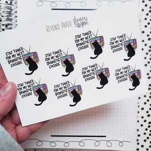May include: Nine black and white stickers with a cartoon cat watching television. The stickers say "Stay tuned for my next dramatic episode".