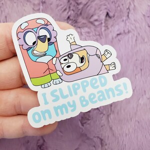 May include: A sticker featuring two cartoon dogs in colorful costumes. The dog on the bottom is lying on its back with its legs in the air. The text on the sticker reads "I slipped on my beans!"