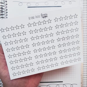 May include: A sheet of white star stickers with the text "BEYOND PAPER flowers" printed at the top. The stickers are arranged in rows and columns, with each star outlined in black. The sheet is held over a notebook with a grid pattern.