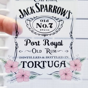 May include: A clear sticker with a black and white design featuring the text "Captain Jack Sparrow's Old No.7 Brand Port Royal Old Rum Distilled & Bottled in Tortuga". The sticker has a floral design with pink flowers and green leaves.