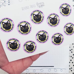 May include: A sheet of stickers with black cats on a pink background. The stickers say "100% Love and Light But Don't Mess With Me".