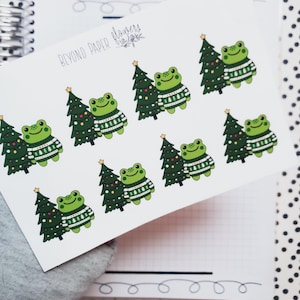 May include: A sheet of stickers featuring green frogs wearing Christmas sweaters and holding Christmas trees. The stickers are on a white background with the text "Beyond Paper Flowers" in the top left corner.