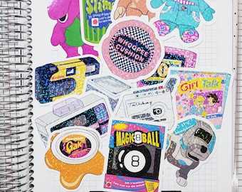Puede incluir: Una colección de pegatinas coloridas en un cuaderno de espiral. Las pegatinas incluyen un dinosaurio morado, un recipiente de slime verde, un mono, un oso azul, una cámara Kodak, una bola mágica 8 y una pegatina que dice "Whoopee Cushion".