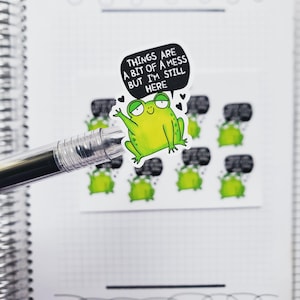 May include: A set of frog stickers with the text "Things are a bit of a mess but I'm still here" on one of the stickers. The frog is green with black eyes and a big smile.