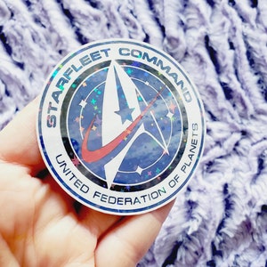 May include: A round sticker with a holographic finish featuring the Starfleet Command logo. The logo is a stylized delta with a red stripe and a blue background. The text "STARFLEET COMMAND" is written in a black arc above the logo, and "UNITED FEDERATION OF PLANETS" is written in a black arc below the logo.