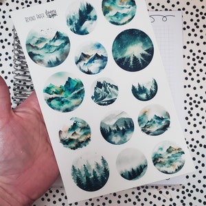 May include: A sheet of 14 watercolor stickers featuring various mountain and forest scenes in shades of blue, green, and white. The stickers are circular and have a white background. The text "Beyond Paper Haven" is printed in the top left corner.