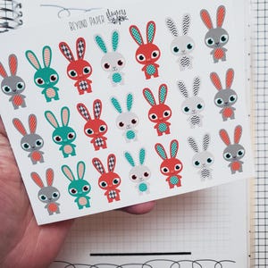May include: Sheet of bunny stickers in red, teal, and gray. Each bunny has a unique pattern on its ears and belly. The stickers are held over a notebook with a grid pattern. The text "BEYOND PAPER flowers" is visible.