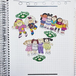 May include: A spiral-bound notebook with several Cabbage Patch Kids stickers. The stickers feature various dolls in colorful outfits, with the Cabbage Patch Kids logo. The notebook has a grid pattern on the pages.