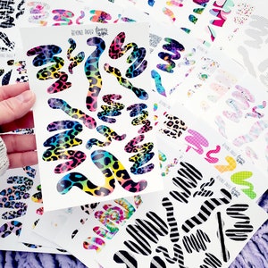 May include: A stack of colorful sticker sheets with various designs, including leopard print, stripes, and tie-dye. The sticker sheets are labeled "Beyond Paper Happy".