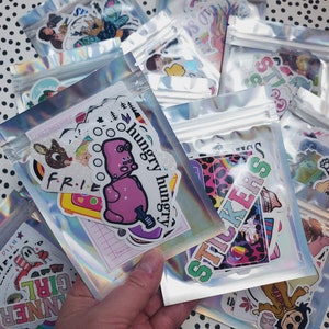 May include: A collection of colorful stickers in various designs, including a pink hippo with the text "hungry hungry", a rainbow unicorn, and a leopard print design with the text "stickers". The stickers are packaged in clear plastic bags with zippers.
