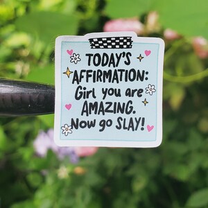 May include: A light blue sticker with the text "TODAY'S AFFIRMATION: Girl you are AMAZING. Now go SLAY!" is decorated with hearts and flowers. The sticker is held in place with black and white checkered tape.