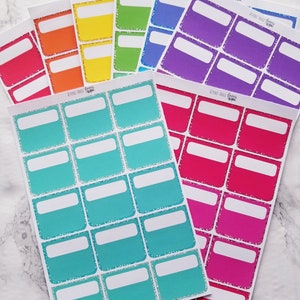 May include: A set of colorful blank label stickers with glitter accents. The stickers are in various colors, including red, orange, yellow, green, blue, purple, and pink. The stickers are all rectangular and have a white border. The text "Beyond Paper Planners" is printed on some of the stickers.