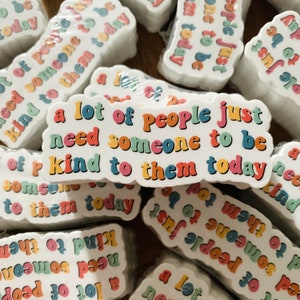 Op de afbeelding: Een stapel kleurrijke stickers met de tekst "a lot of people just need someone to be kind to them today" erop gedrukt. De stickers zijn wit met regenboogkleurige letters.